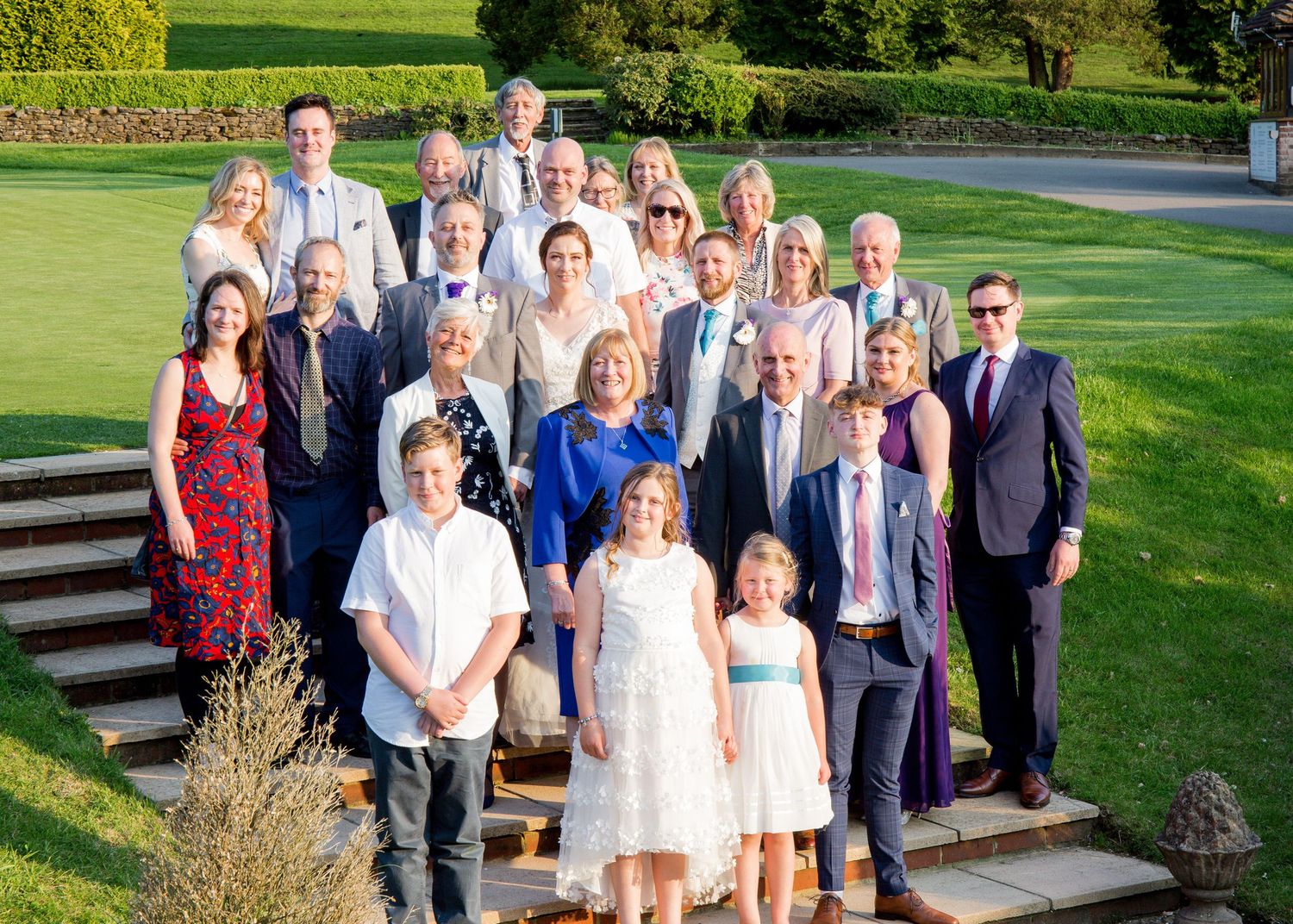Large wedding group photo of guests posing on outdoor steps surrounded by lush green gardens.