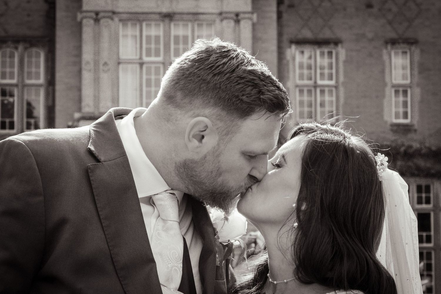 Black and white wedding photo of bride and groom sharing a romantic kiss outside a historic brick building.