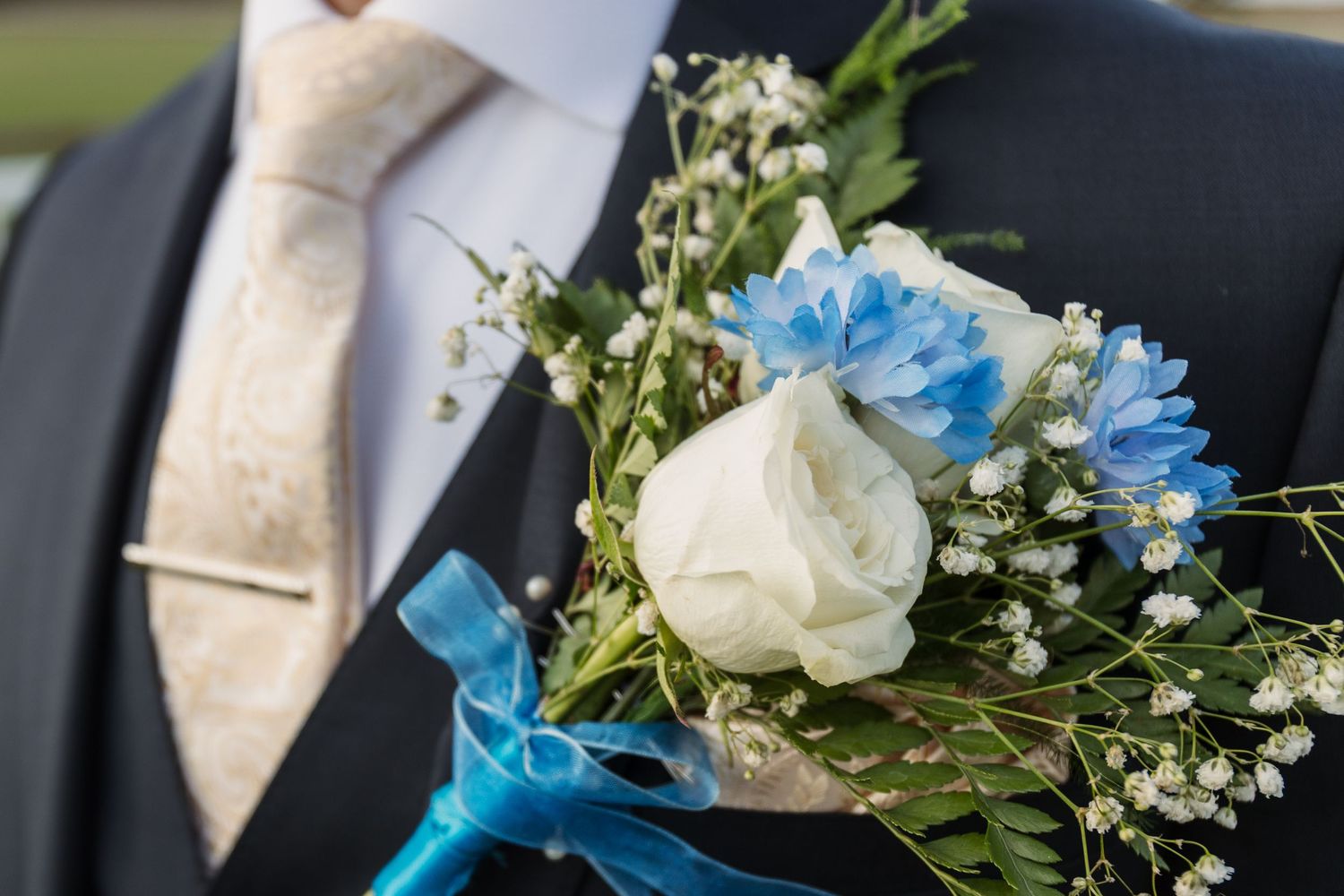 Close-up of a wedding boutonniere with white rose, blue flowers, and greenery on a groom's suit with beige tie.