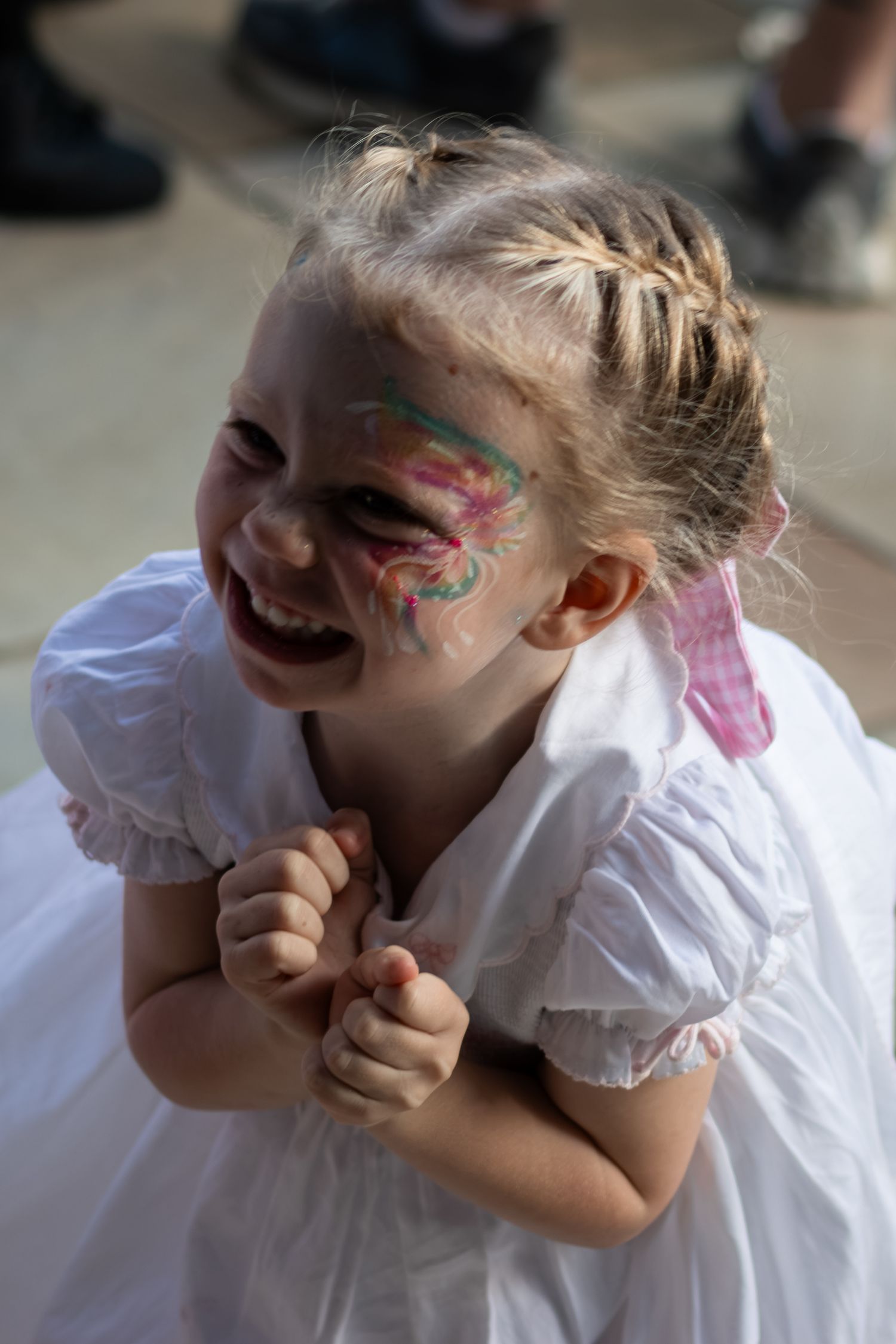 Laughing little girl with face paint and braided hair wearing a white dress at an outdoor event.