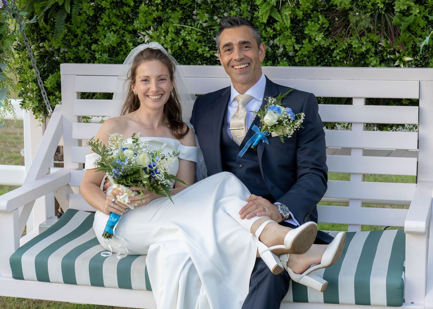 Happy bride and groom sitting on a white garden bench, bride holding a blue and white bouquet on their wedding day.