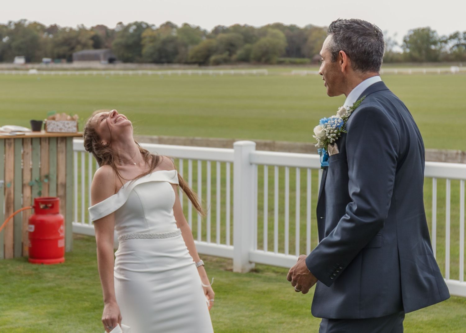 A bride in a white off-shoulder dress laughs joyfully beside a groom in a navy suit at an outdoor wedding venue.