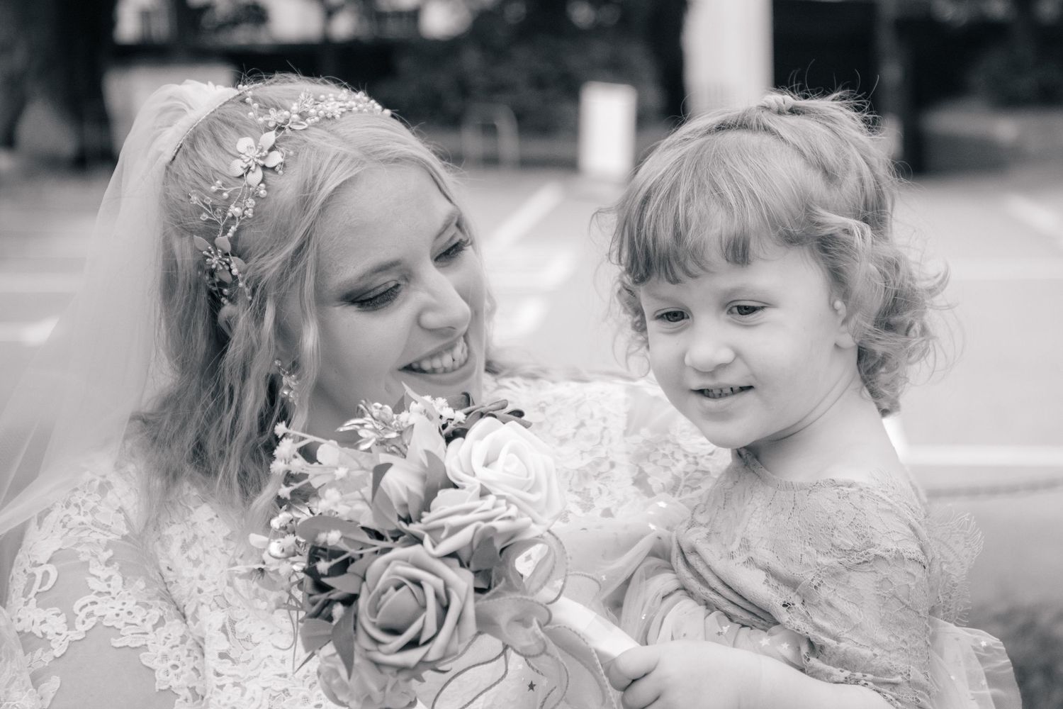 Bride in lace wedding dress holding bouquet smiling at young flower girl in black and white photo.