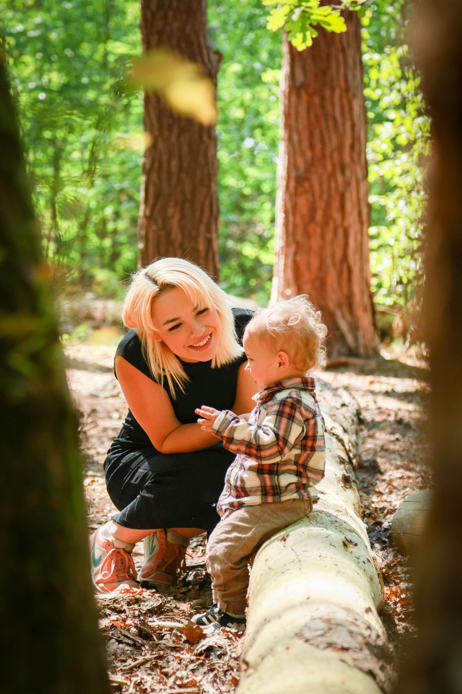 Smiling blonde mother crouching with toddler boy by a fallen log in a sunny green forest.