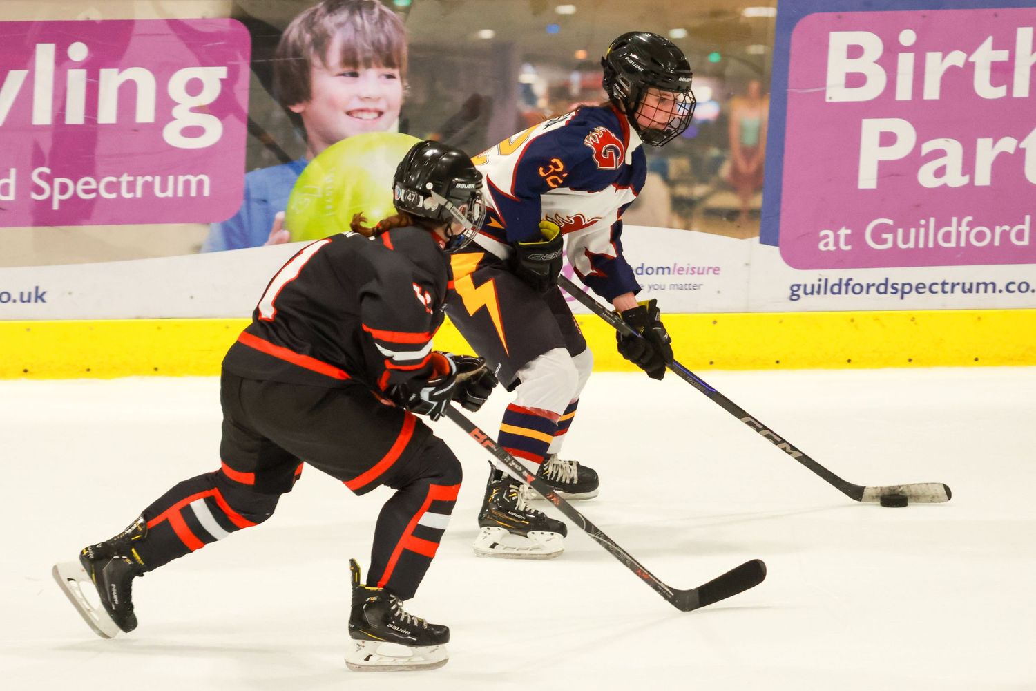 Two young female ice hockey players battle for puck on ice with Guildford Spectrum birthday parties banner in background.