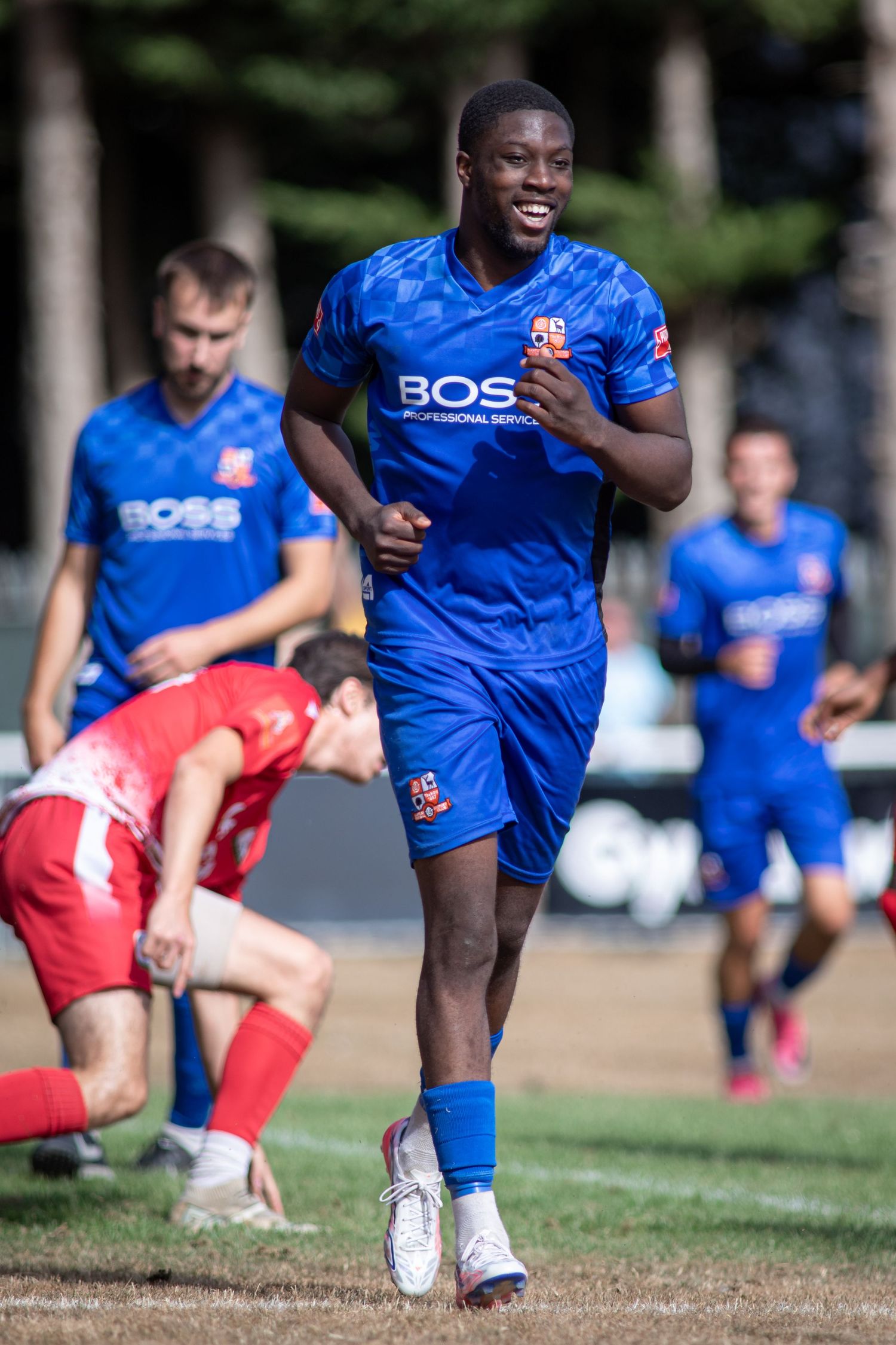 Blue team footballer in BOSS jersey runs past fallen opponent during outdoor soccer match.