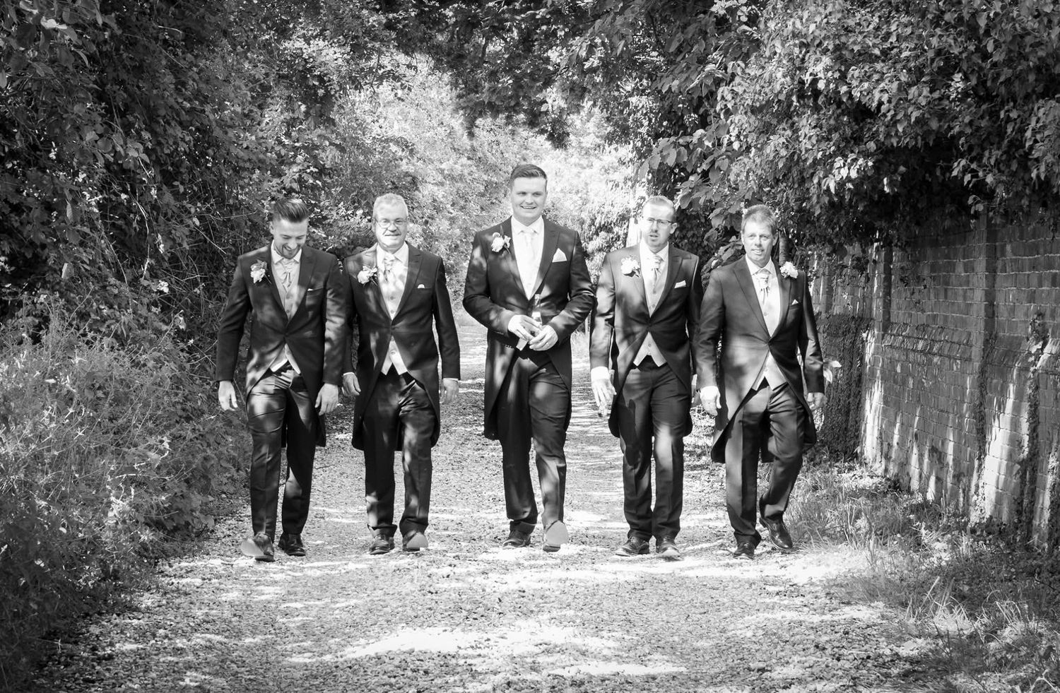 Black and white photo of five groomsmen in formal suits walking confidently down a tree-lined path.