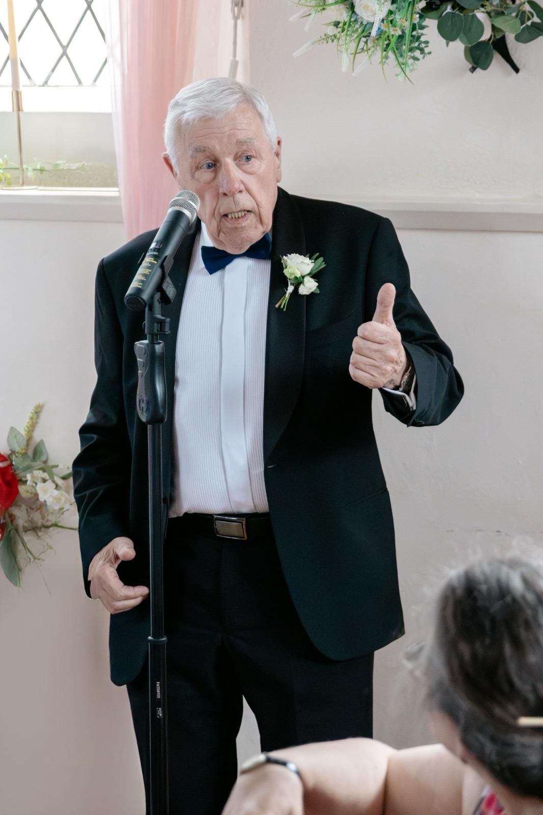 Elderly man in tuxedo with boutonniere giving thumbs up while speaking at microphone during wedding reception.