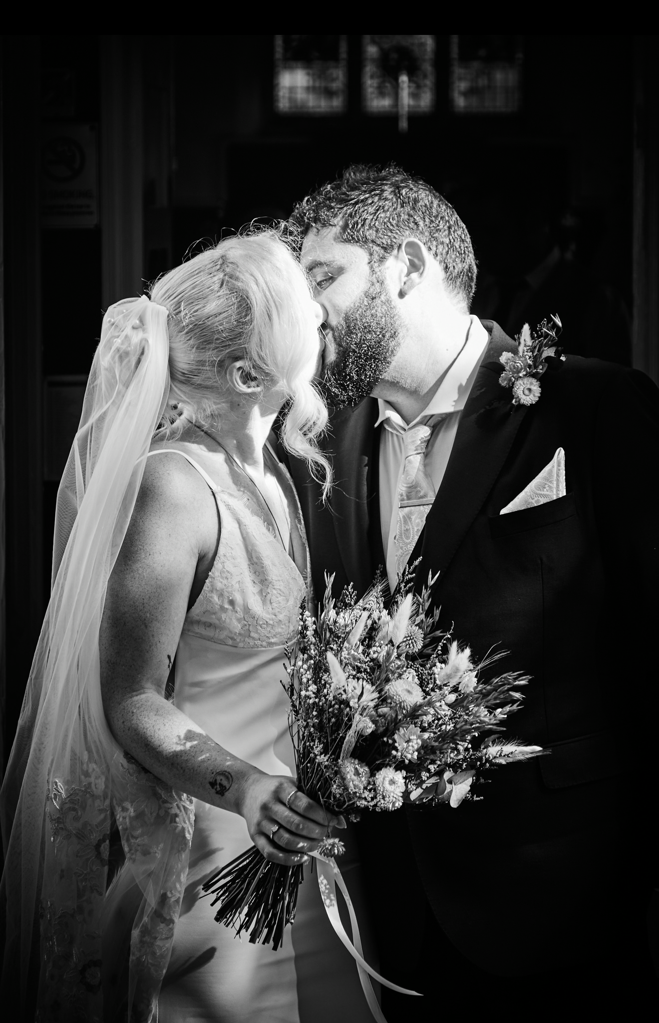 Black and white wedding photo of couple sharing romantic kiss, bride in lace gown holding floral bouquet.