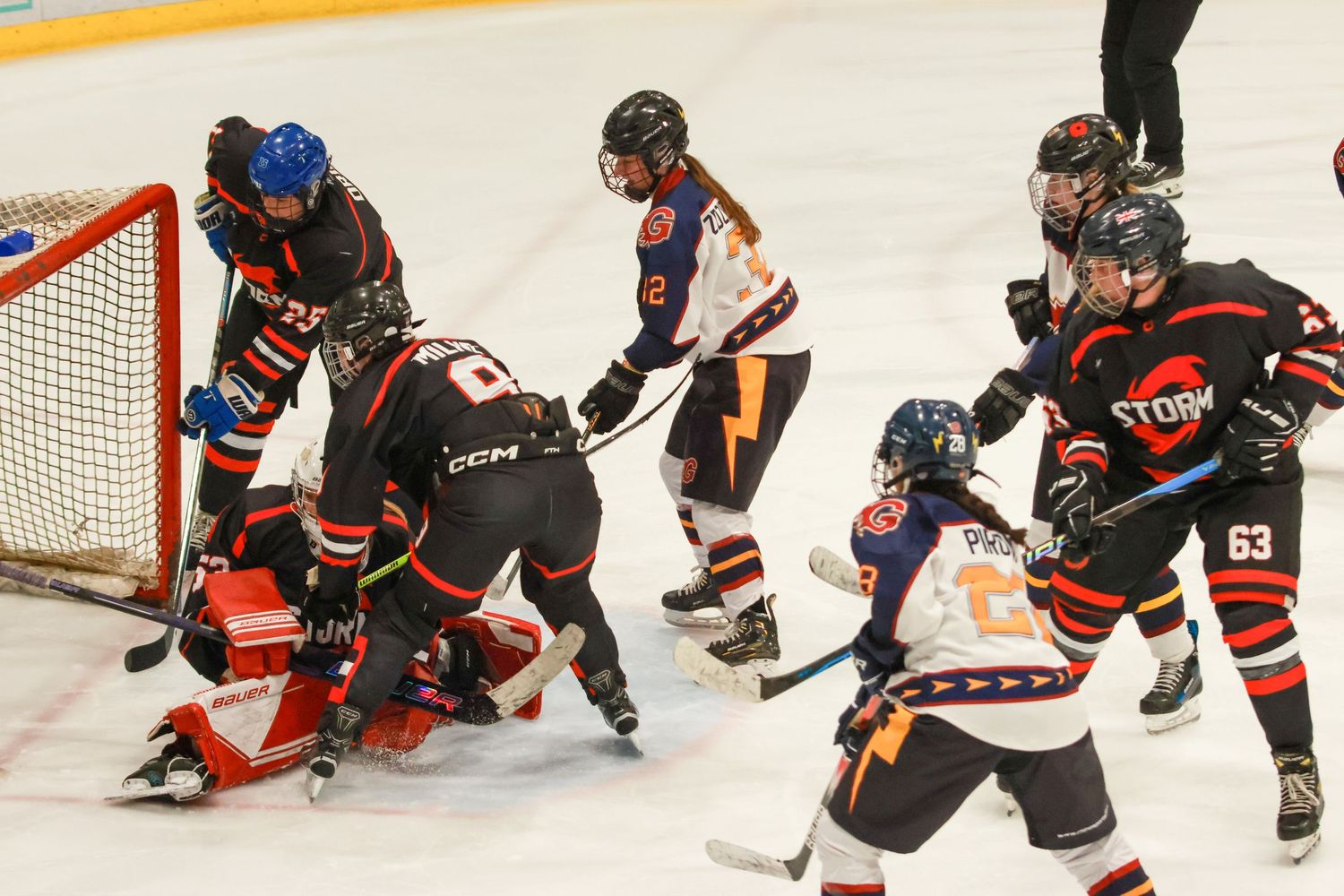 Intense ice hockey scramble in front of goal with Storm goalie down as multiple players from both teams battle for puck.