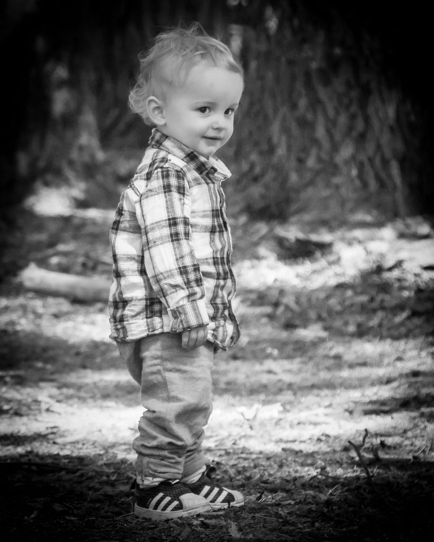 Black and white portrait of a toddler boy in plaid shirt and Adidas sneakers smiling outdoors.