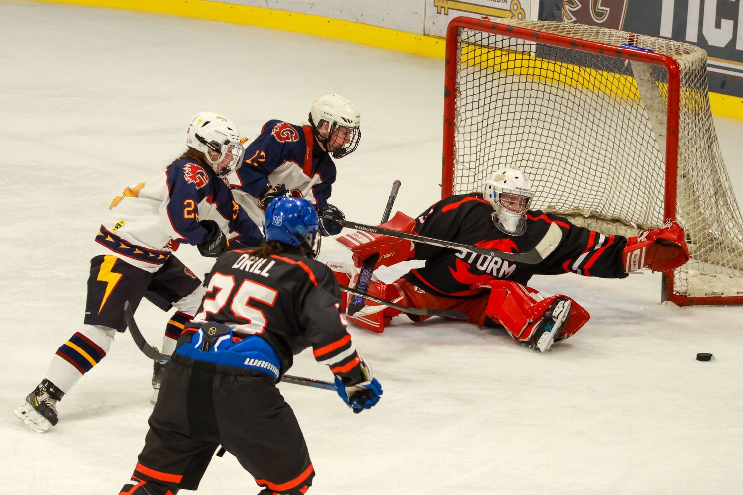 Ice hockey players crash the net as goalie makes save during intense game action with player Hill number 25 visible.