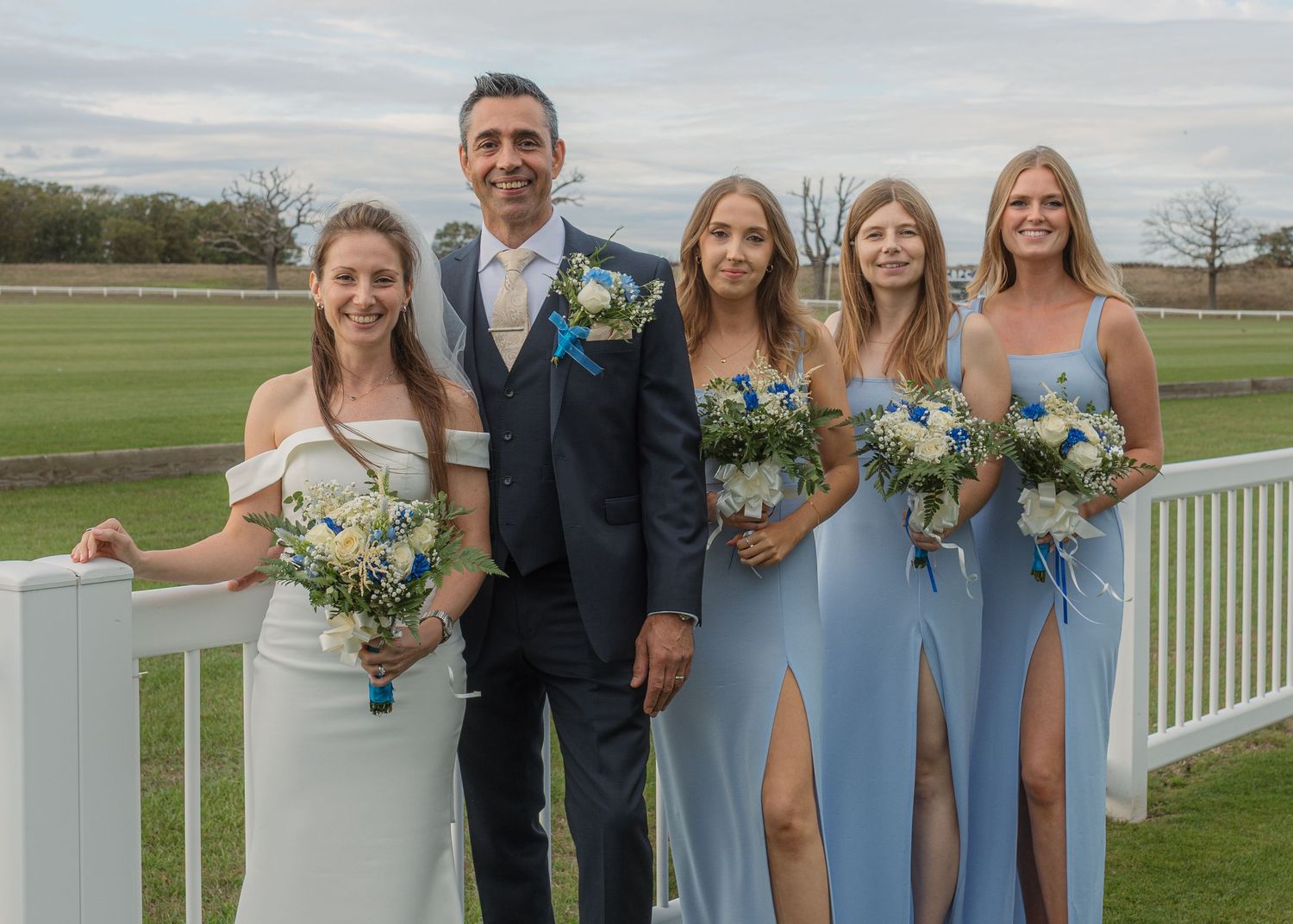 Wedding party posing outdoors, bride in white, groom in suit, three bridesmaids in blue dresses holding floral bouquets.