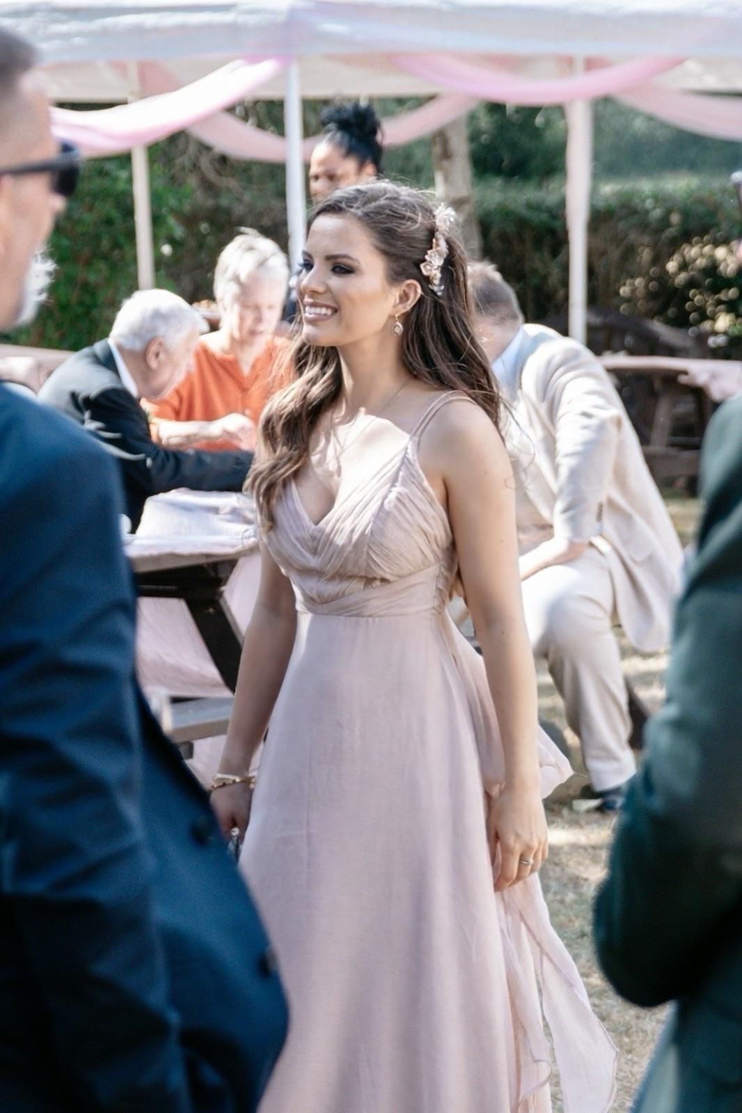Smiling bridesmaid in elegant pink gown at outdoor wedding reception with guests in background.