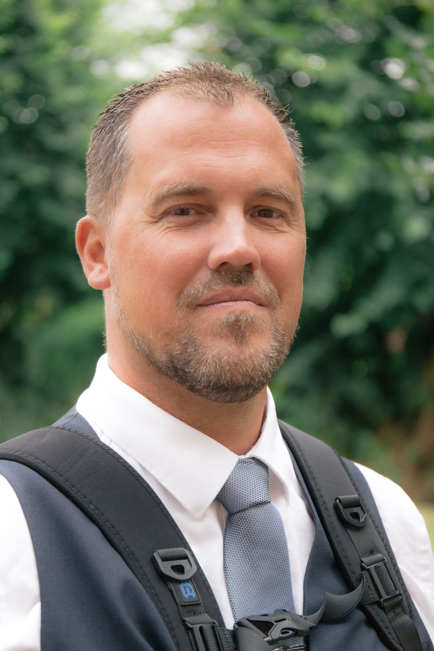 Professional man in white shirt, gray tie and vest smiling outdoors with green foliage background.