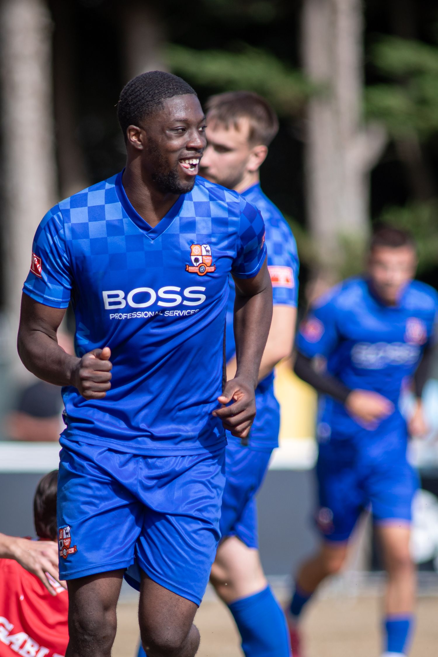 Joyful soccer player in blue BOSS checkered kit celebrates goal with teammates behind him.