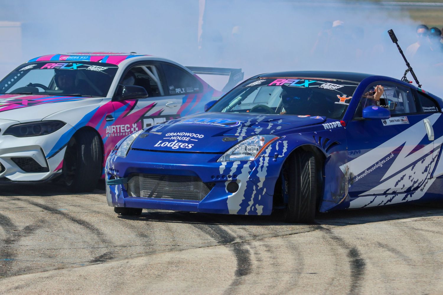 Blue Nissan 350Z drift car with Lodges sponsorship producing smoke alongside a pink BMW.