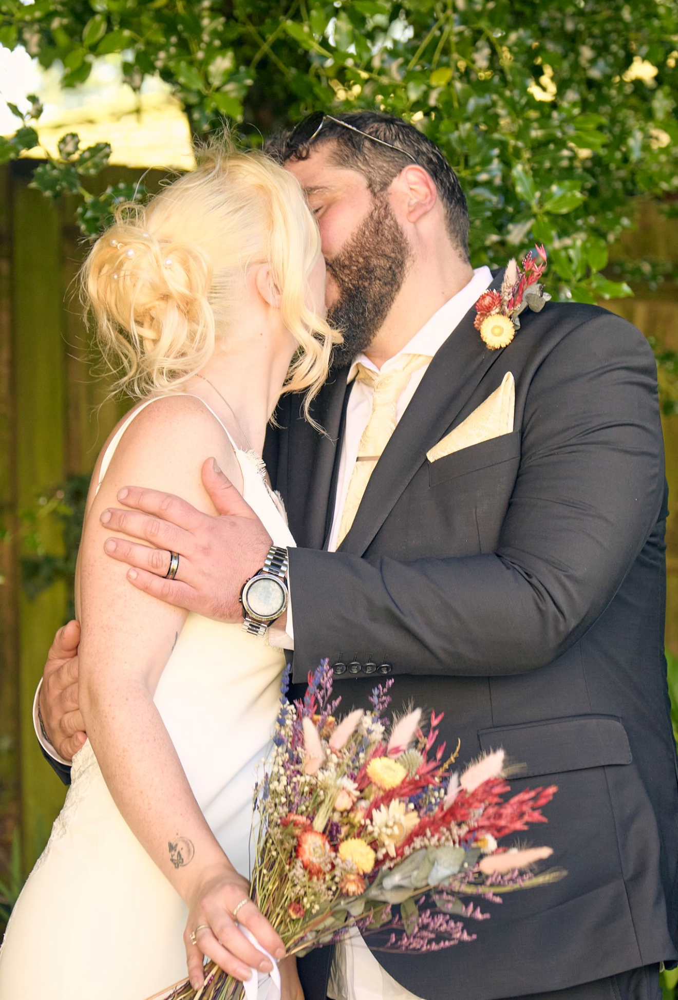 Bride and groom kissing outdoors, bride holding colorful dried flower bouquet, groom in dark suit with boutonniere.