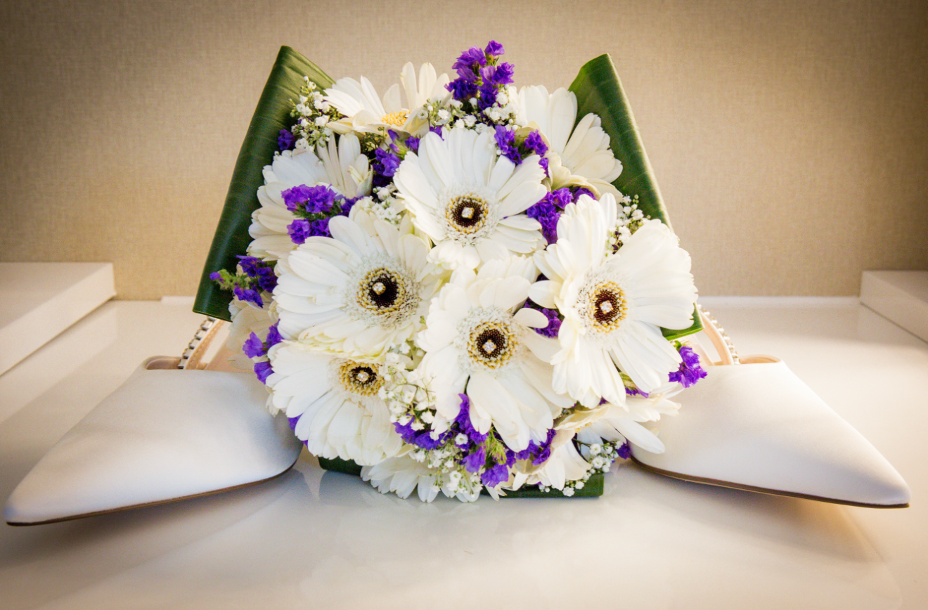 Bridal bouquet of white gerbera daisies with purple accents beside white wedding heels.