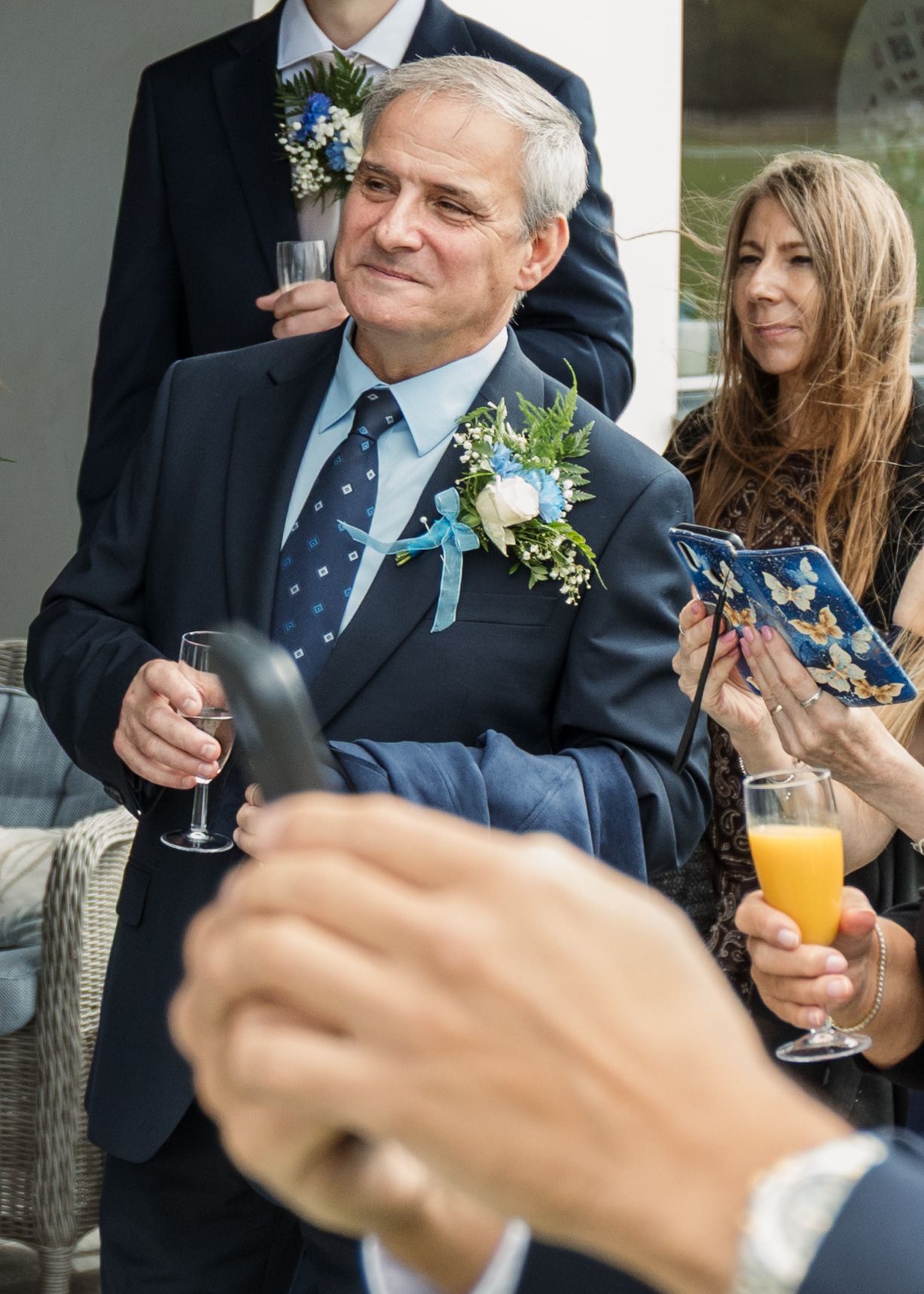 Older man in navy suit with floral boutonniere holding champagne glass at a wedding reception.