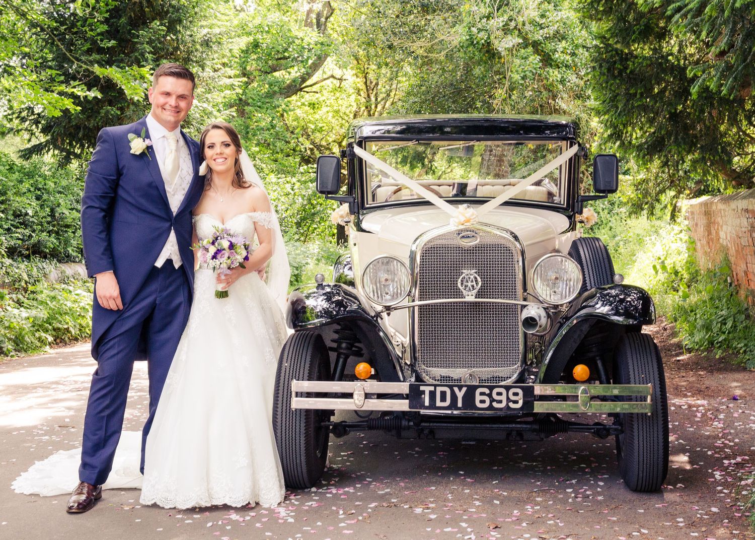 Bride and groom posing beside a vintage black and cream wedding car with white ribbons on a tree-lined driveway.