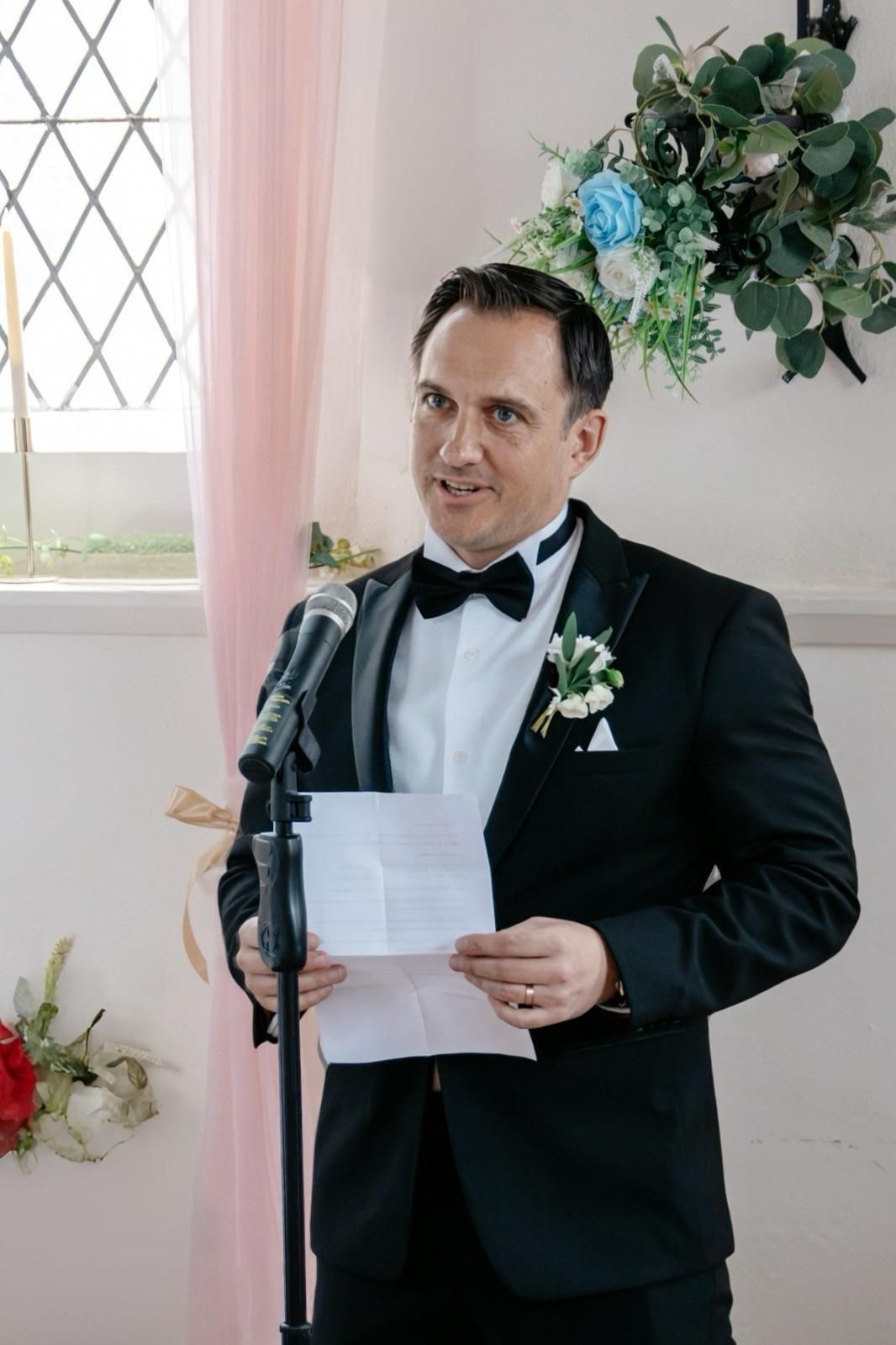 Groom in black tuxedo with bow tie reads vows at microphone stand during wedding ceremony.