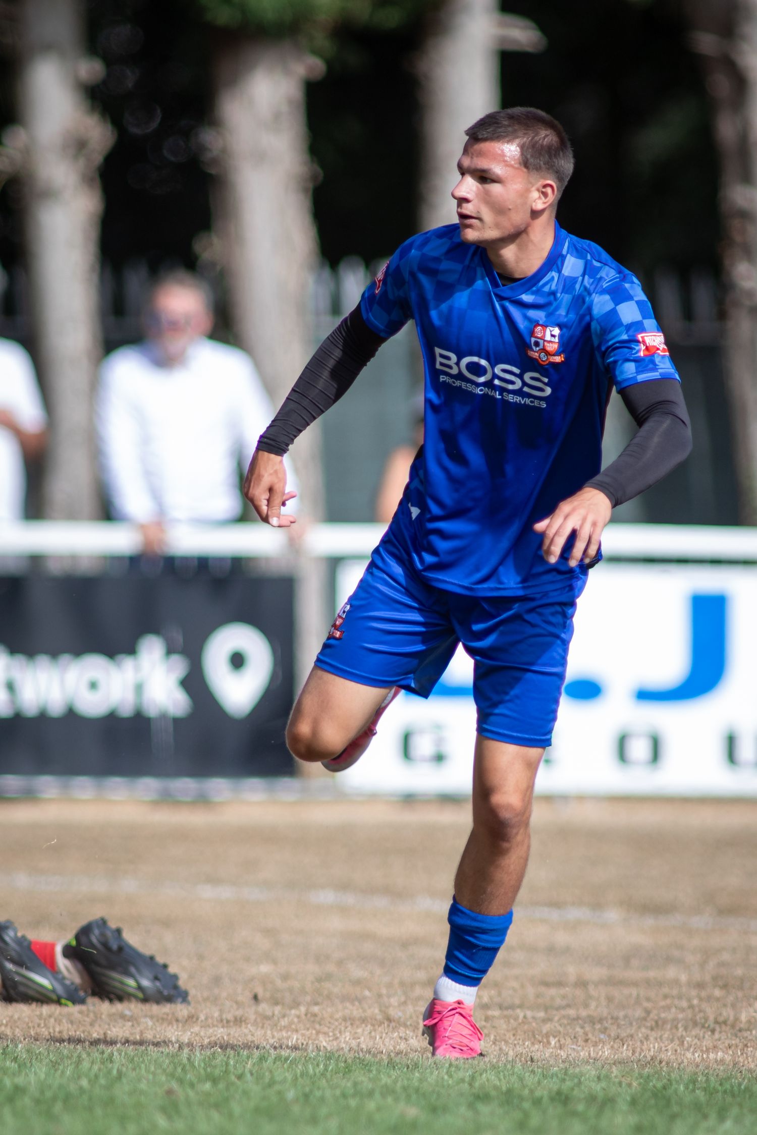 Blue BOSS sponsored footballer in checkered kit runs during sunny outdoor soccer match.