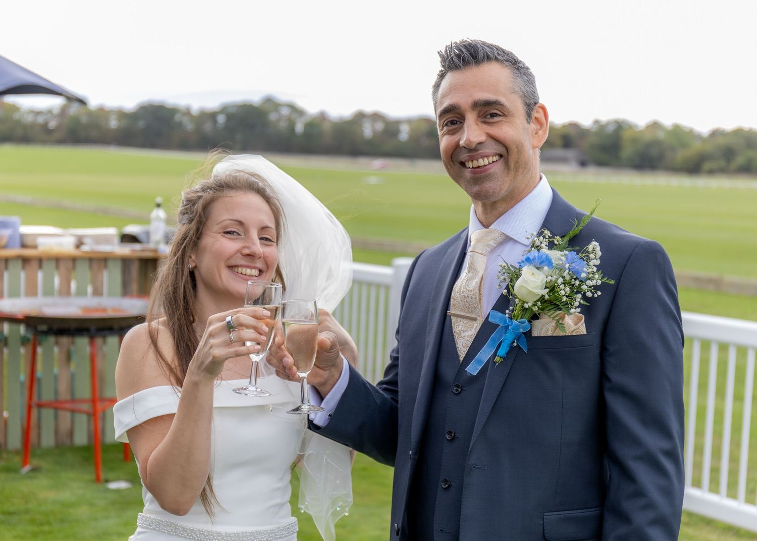 Bride and groom toasting with champagne glasses at outdoor wedding venue, wearing white dress and navy suit.