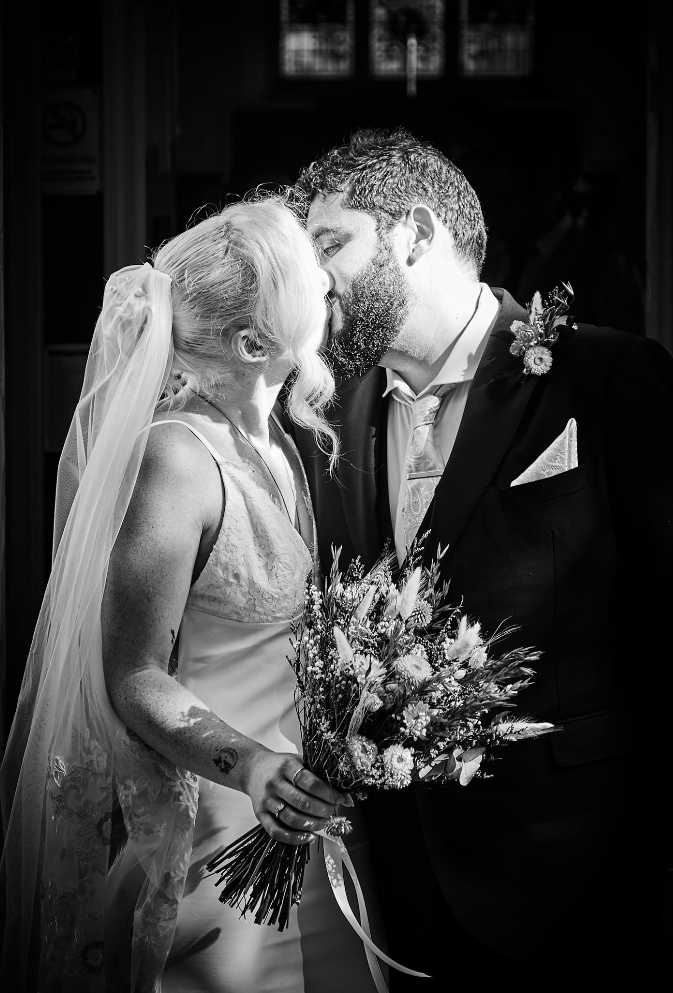 Black and white photo of bride and groom sharing first kiss, bride holding wildflower bouquet wearing veil.