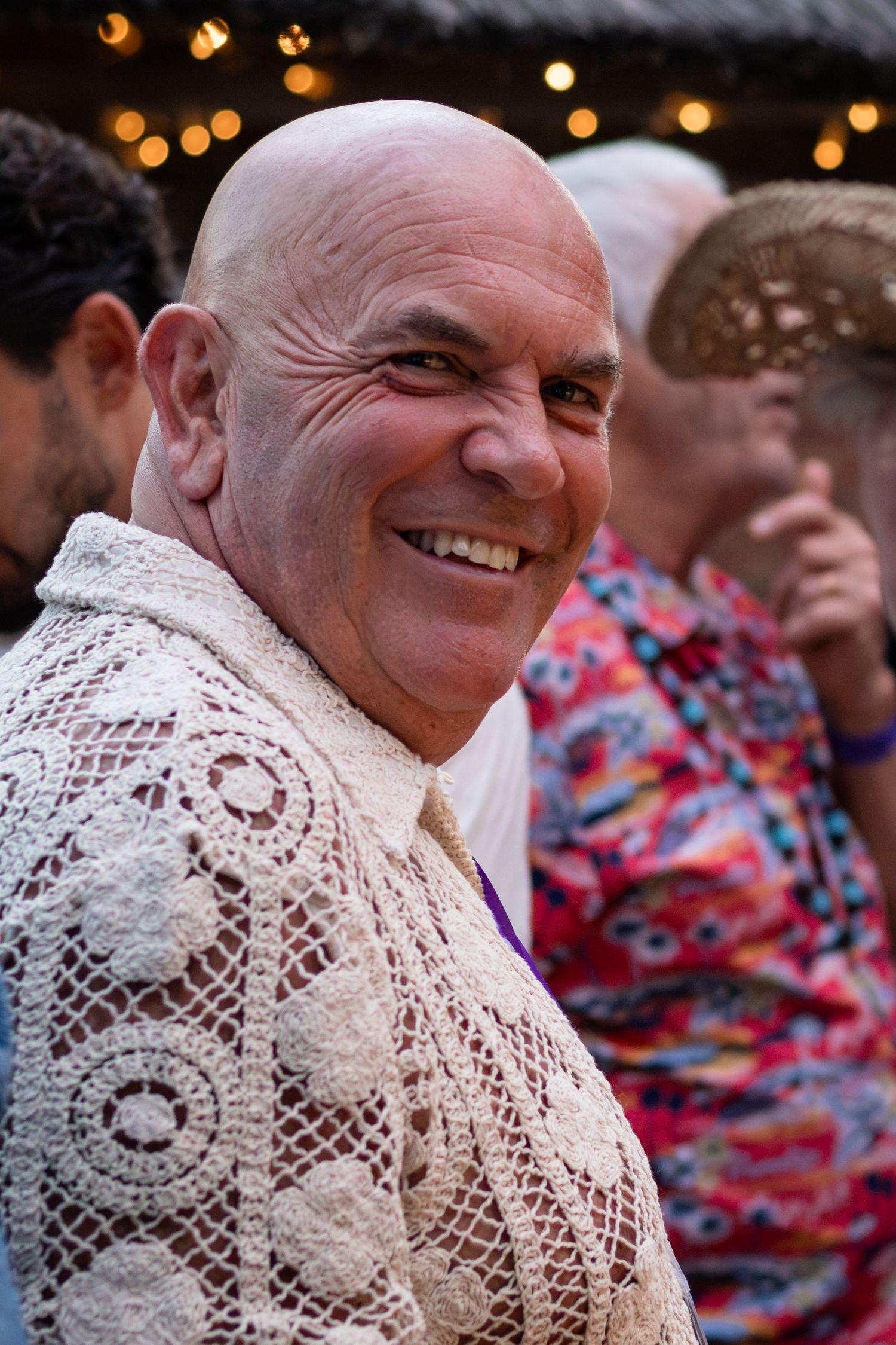 A bald man smiling warmly wearing a lace shirt at an outdoor party.