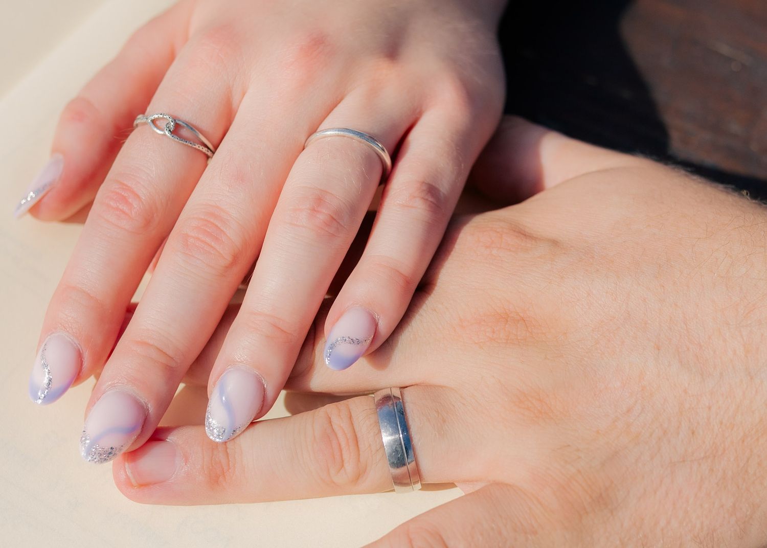 Two hands with matching silver wedding bands and lavender almond-shaped nails rest together, symbolizing love and commitment.