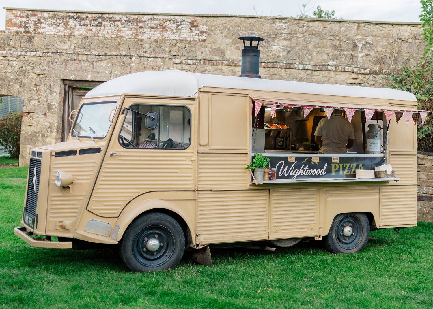 Vintage beige Citroen H Van food truck with bunting and serving hatch, parked on grass near stone wall.