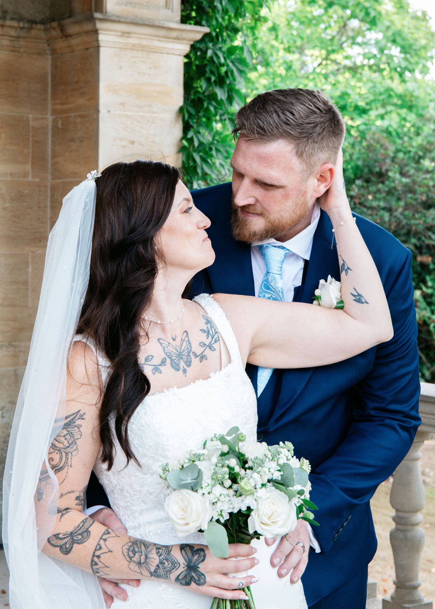 Tattooed bride in white gown holds floral bouquet while groom in navy suit tenderly gazes at her outdoors.