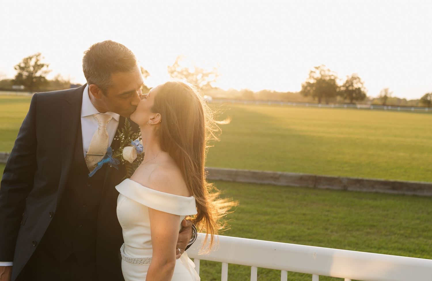 Bride and groom sharing a romantic sunset kiss on a golf course, bride in off-shoulder gown, groom in navy suit.