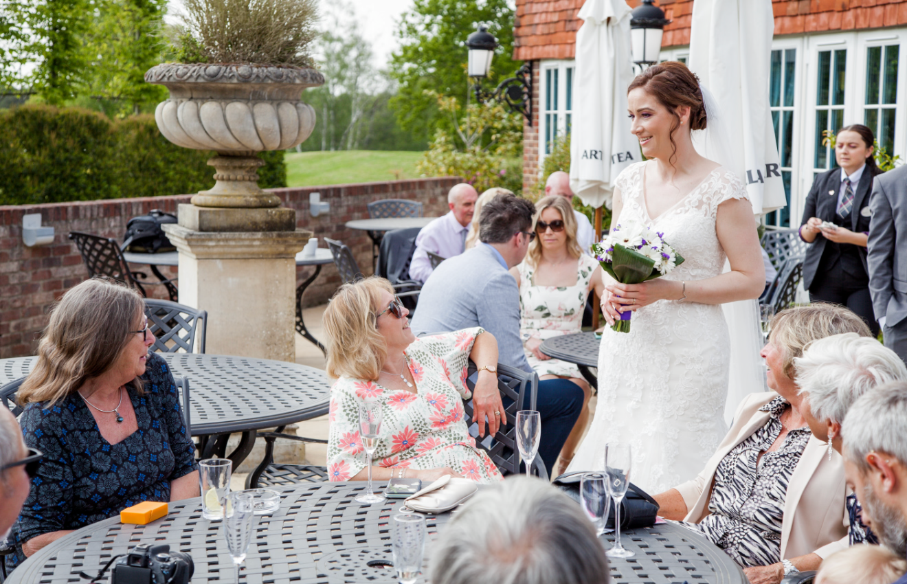 Bride in white gown holding bouquet greeting seated wedding guests at an elegant outdoor garden reception.