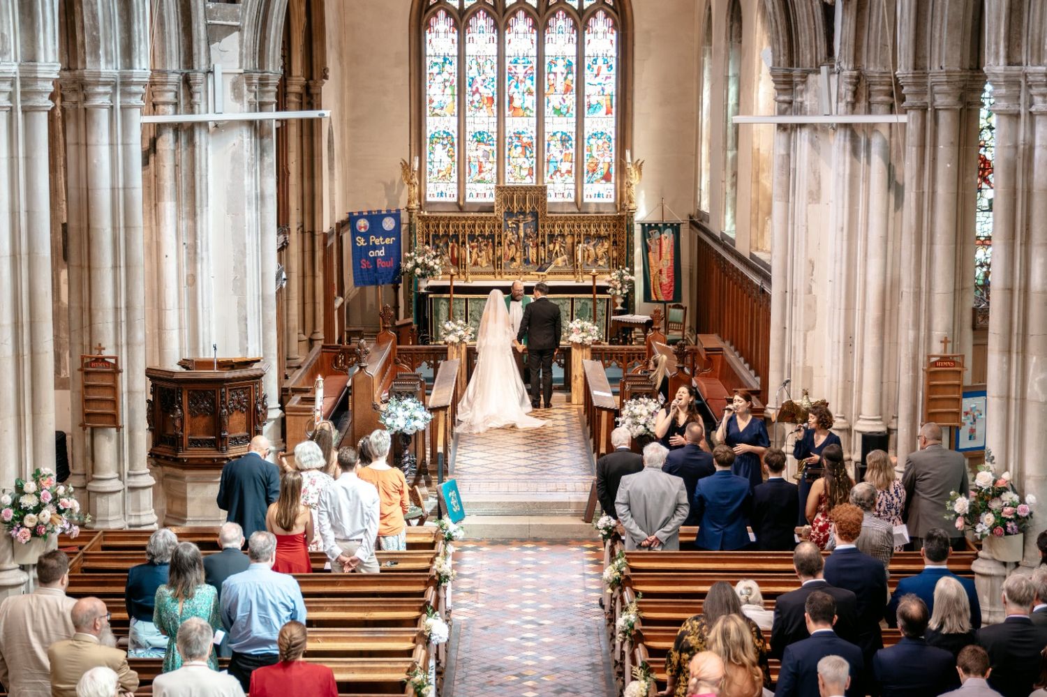 Guests seated in ornate church pews during a wedding ceremony with stained glass windows and floral arrangements.