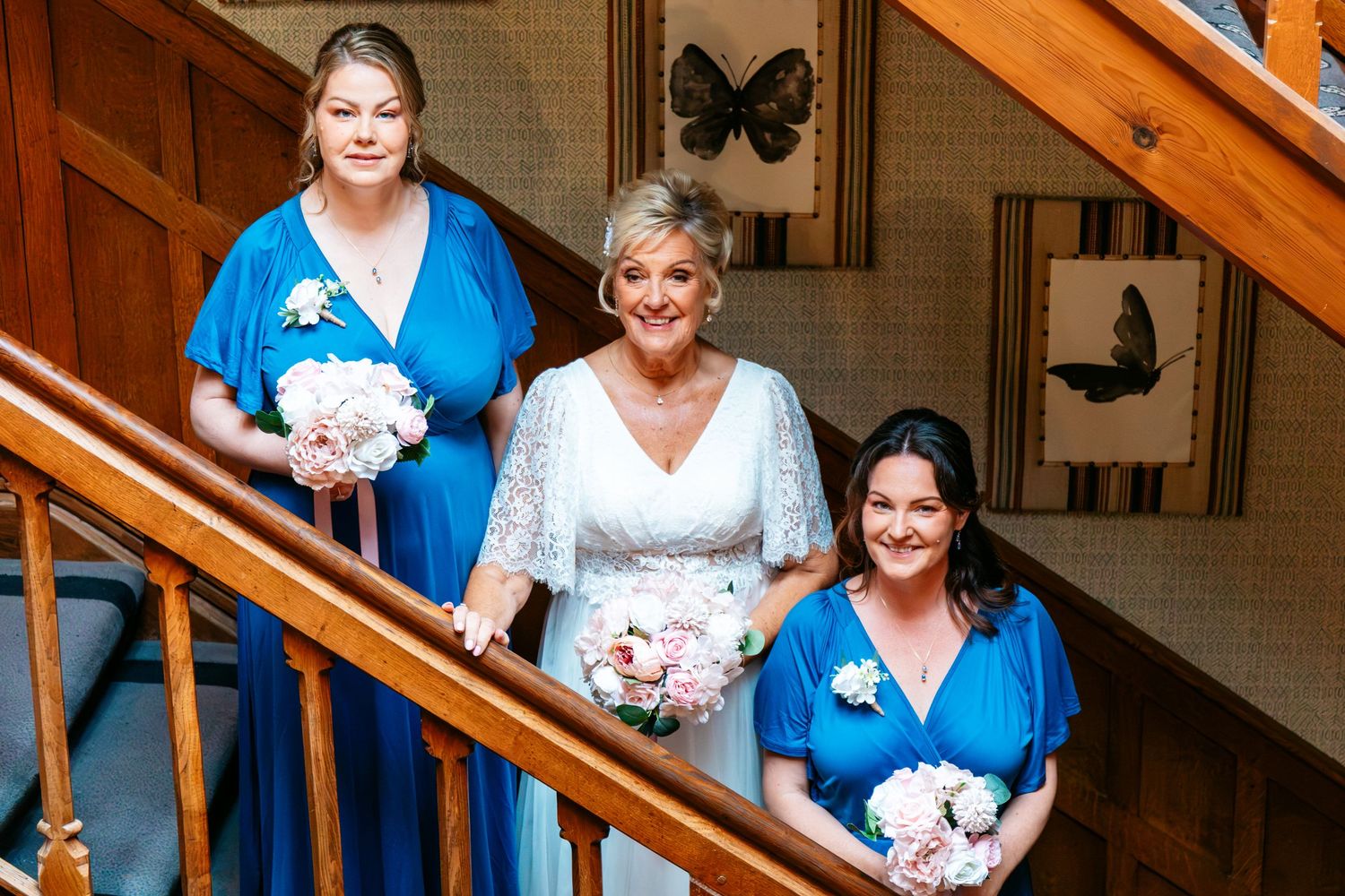 Bride in white lace dress with two bridesmaids in blue gowns holding pink bouquets on a wooden staircase.