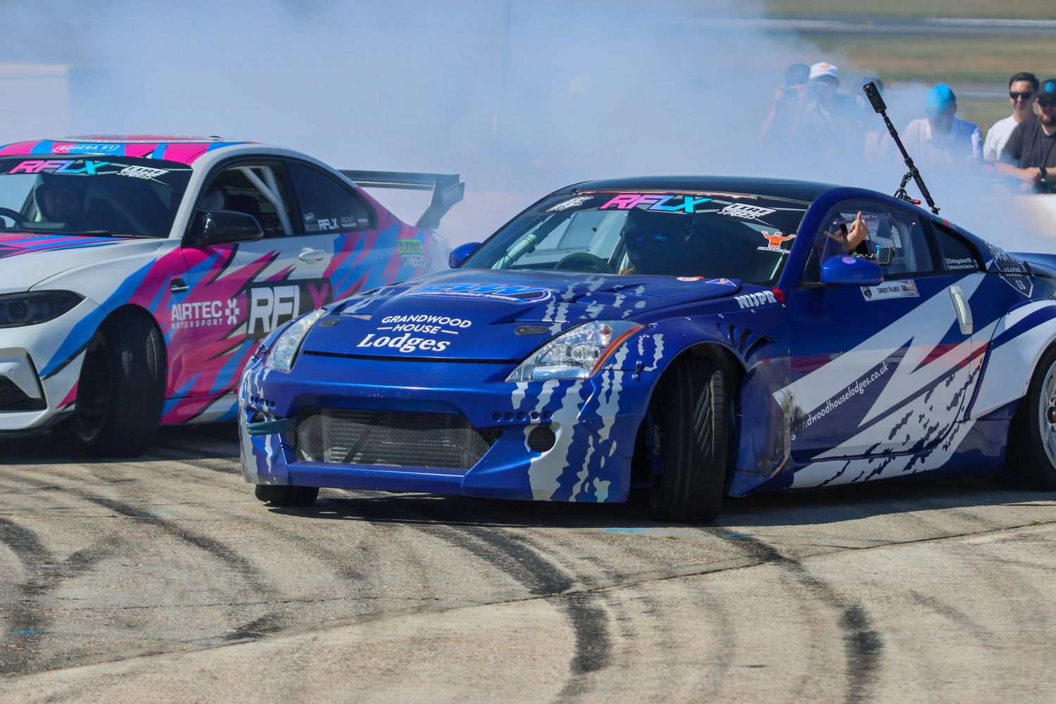 Blue Nissan 350Z drift car and pink BMW M2 producing smoke during a tandem drift battle.