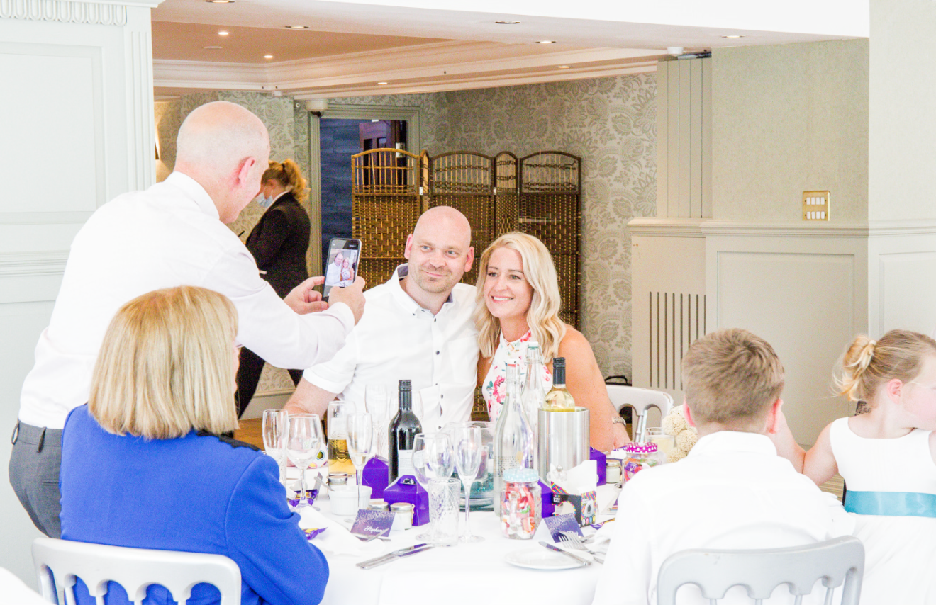 Wedding guests celebrating at reception table, raising glasses in a toast with bottles on table.