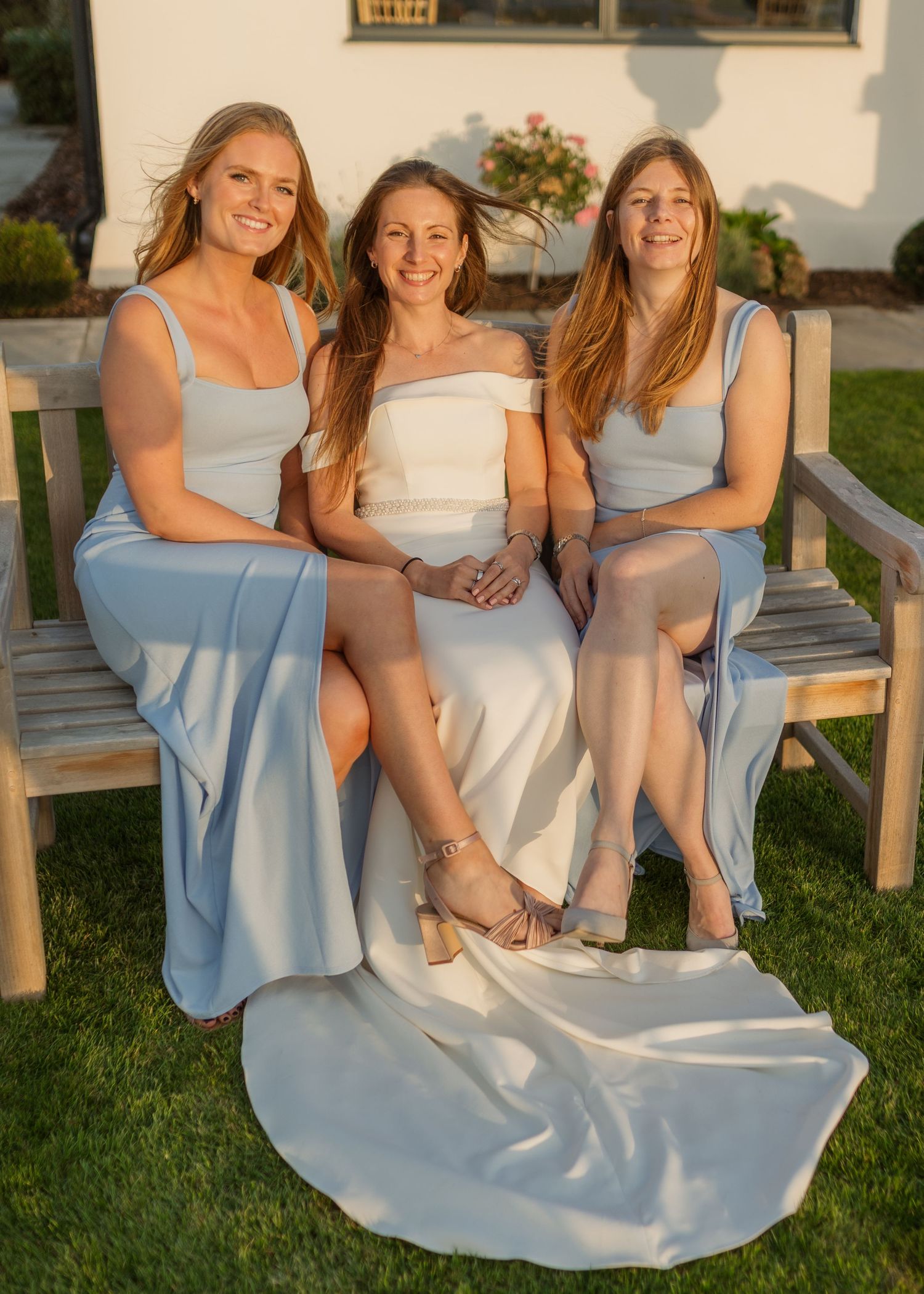 Bride in white gown sits with two bridesmaids in light blue dresses on a wooden bench outdoors.