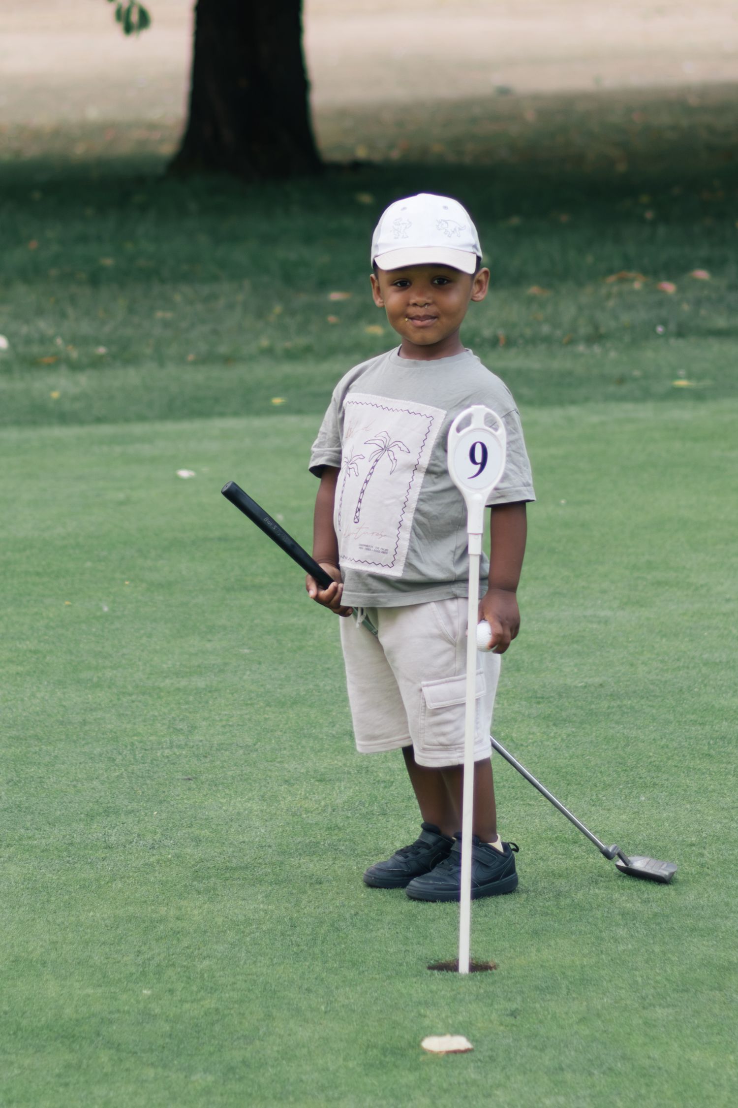 Young boy in white cap and khaki shorts holding golf club standing on green golf course.