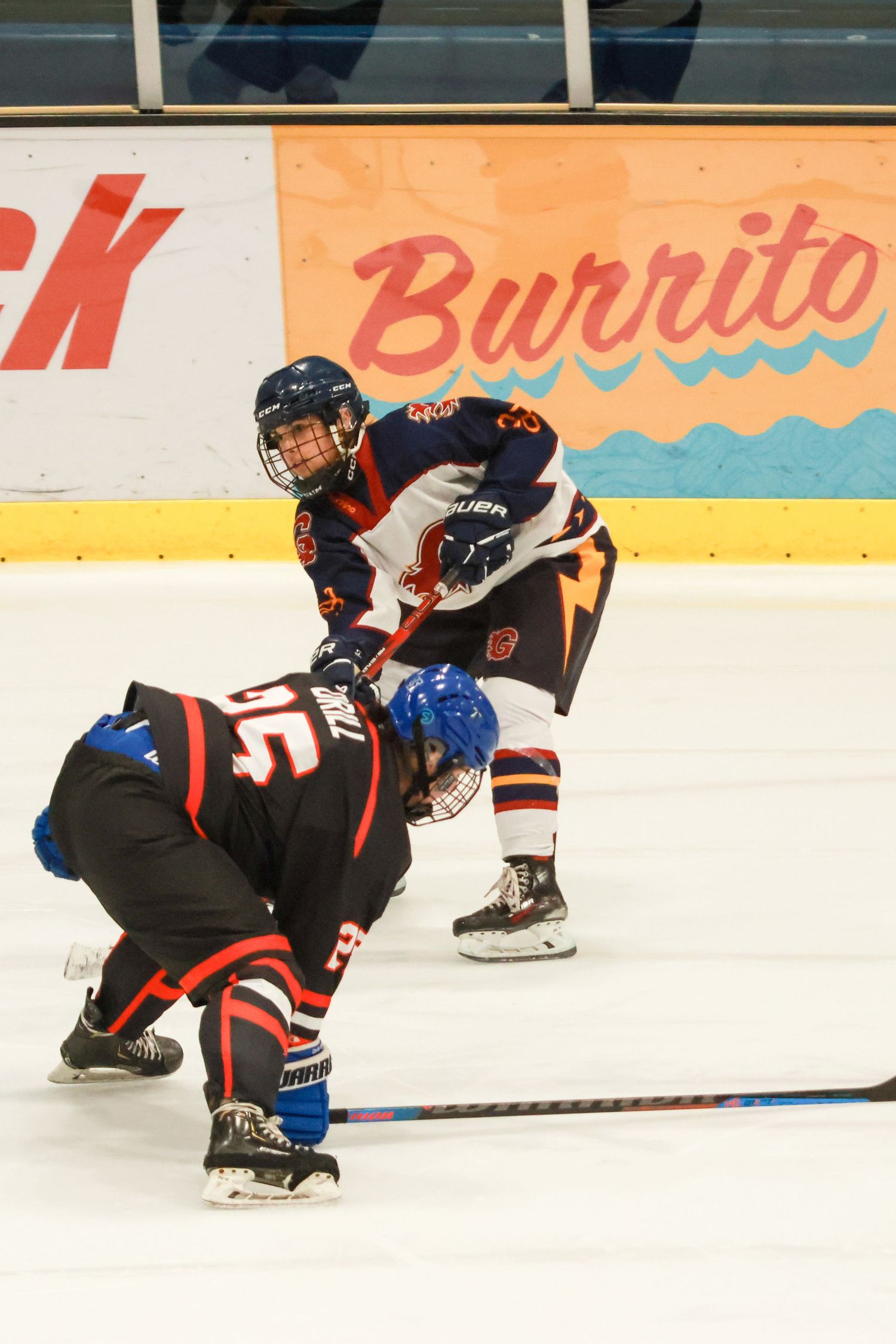 Two youth ice hockey players compete for the puck near a Burrito advertisement board on the rink.