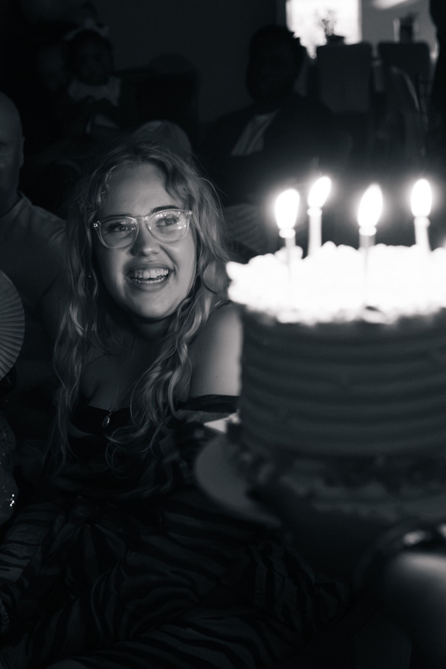 Smiling young woman with glasses celebrates birthday with lit candle cake in black and white photo.
