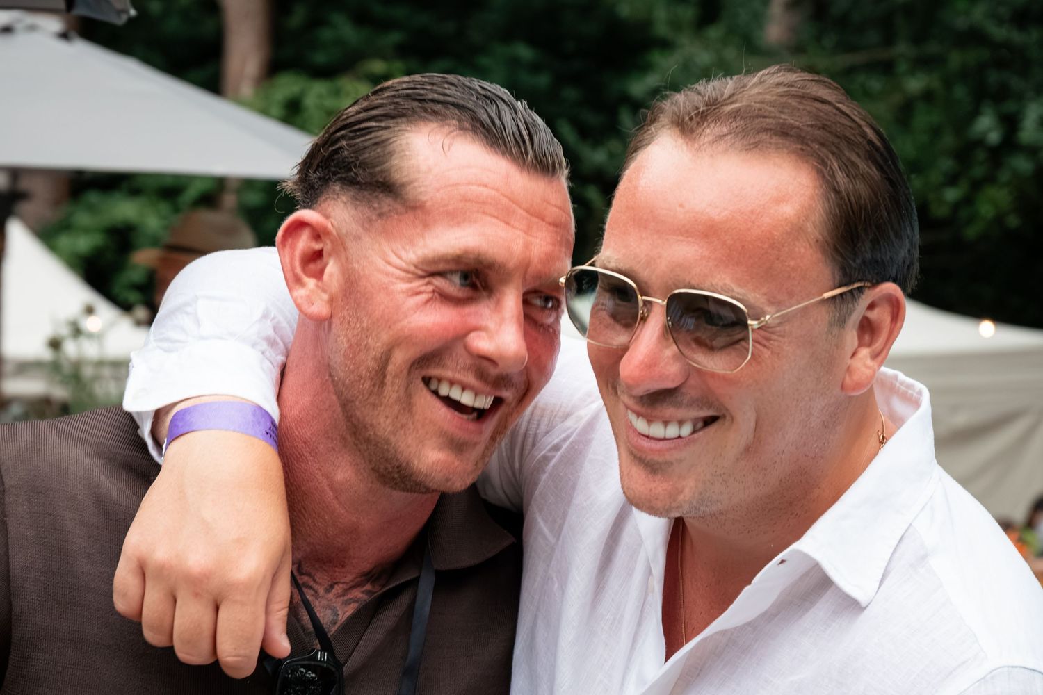 Two smiling men in sunglasses share a friendly embrace at an outdoor social gathering.