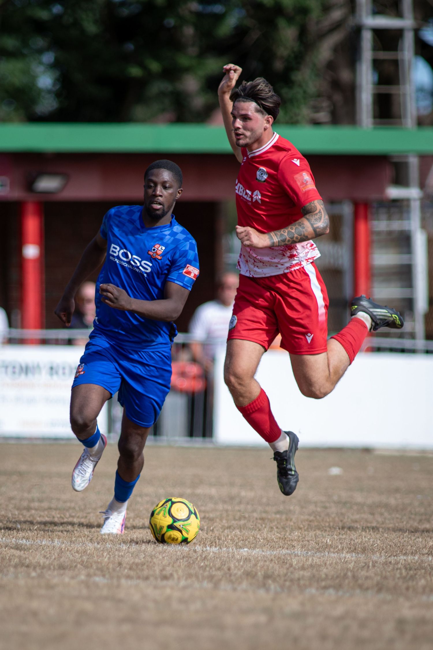Two soccer players compete for the ball, one in red and one in blue BOSS kit, on a dry grass pitch.