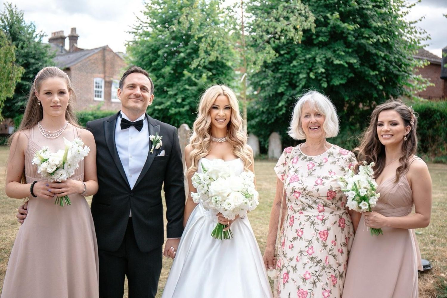 Bride in white gown poses with groom in tuxedo and bridesmaids in blush dresses holding bouquets at outdoor wedding.
