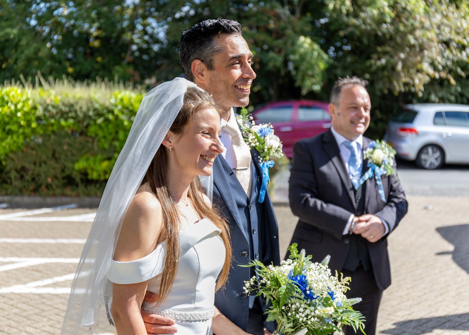 Happy bride and groom smiling outdoors on their wedding day, bride holding blue and white floral bouquet wearing veil.