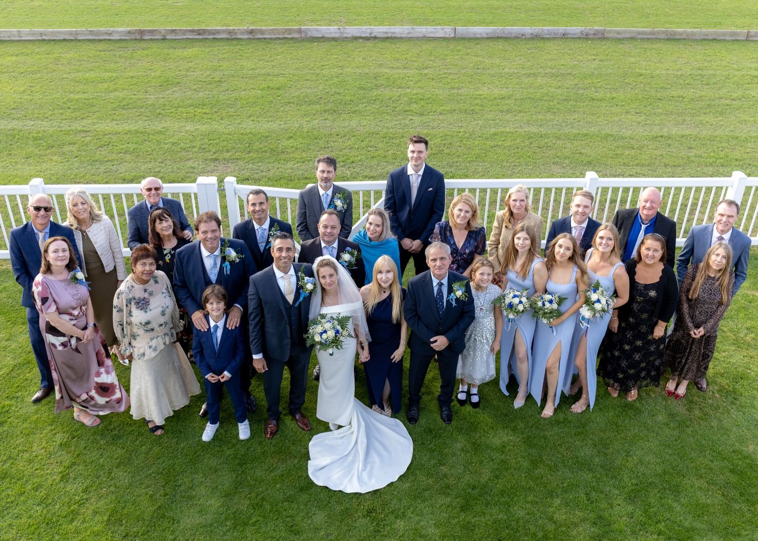 Large wedding group photo outdoors on green grass, bride in white gown surrounded by family and friends in formal attire.