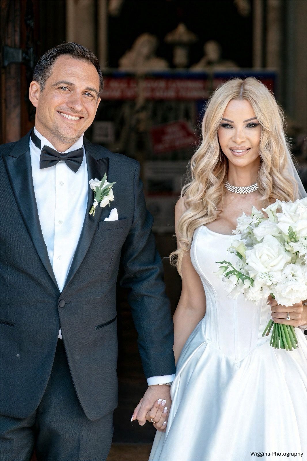 Elegant bride and groom holding hands outside church, bride holding white floral bouquet in satin gown.
