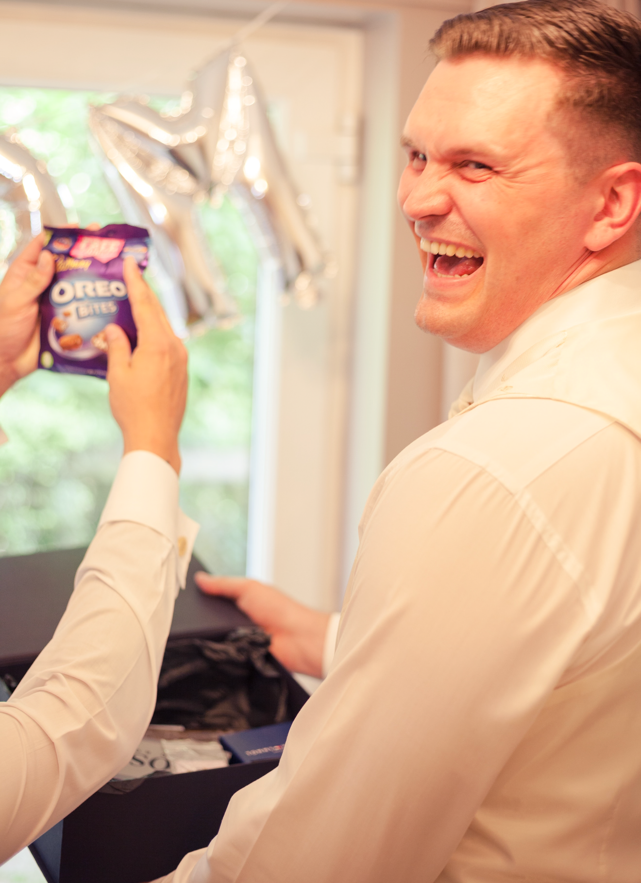 A laughing man in a white shirt receives a pack of Oreo Bites at a celebration with silver balloon decorations.